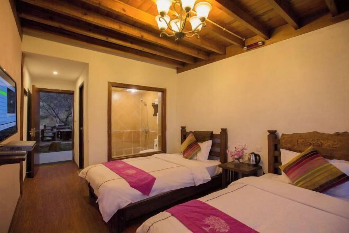 A hotel room with two beds, wooden beams on the ceiling, a TV, and a bathroom visible through a glass door.