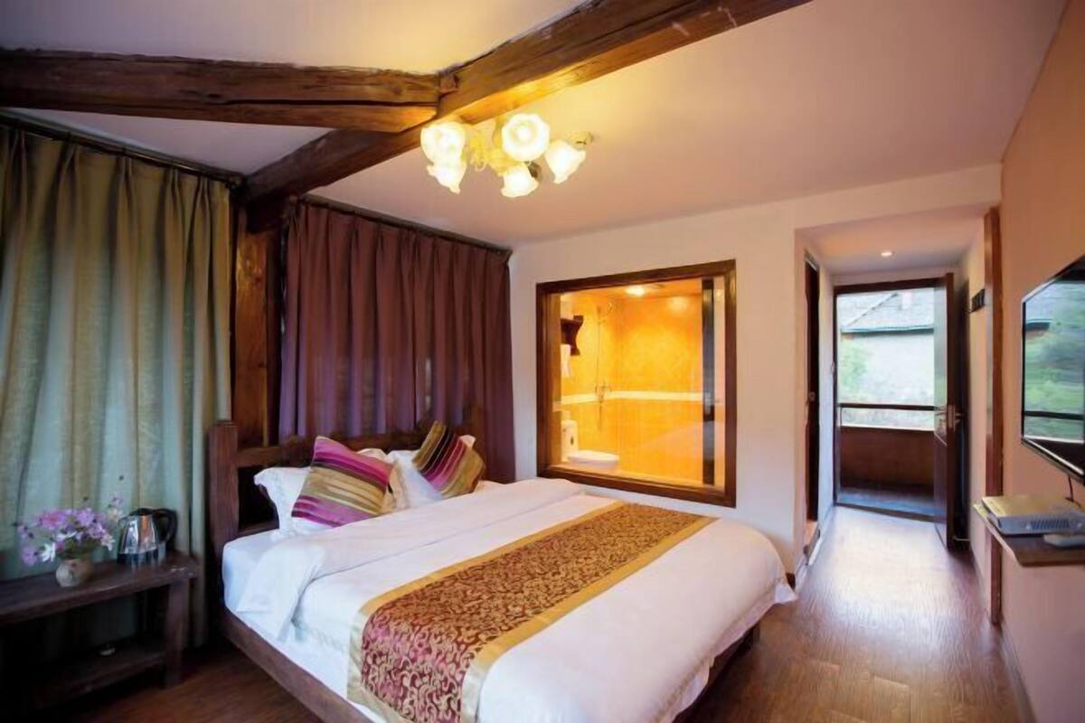 A hotel room with a wooden ceiling, a large bed with multiple pillows, a bedside table with a lamp, and a window with curtains.
