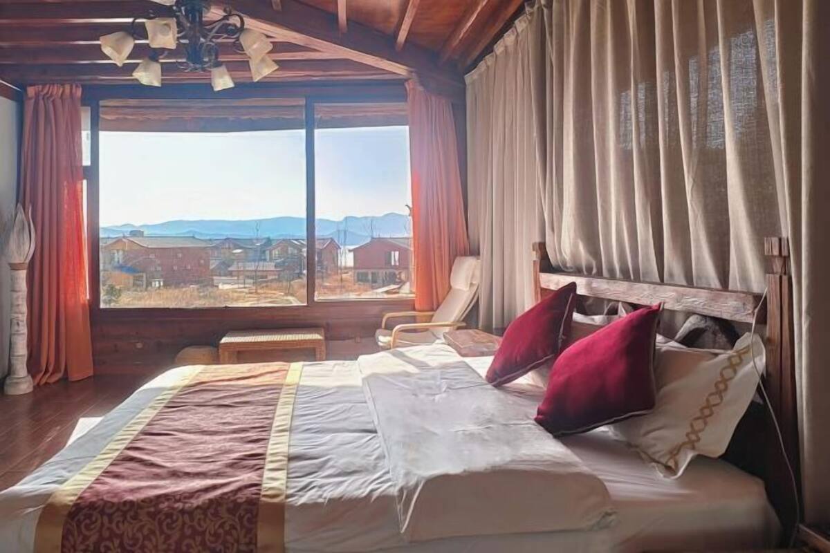 A bedroom with a large bed, a view of houses, and a wooden ceiling.