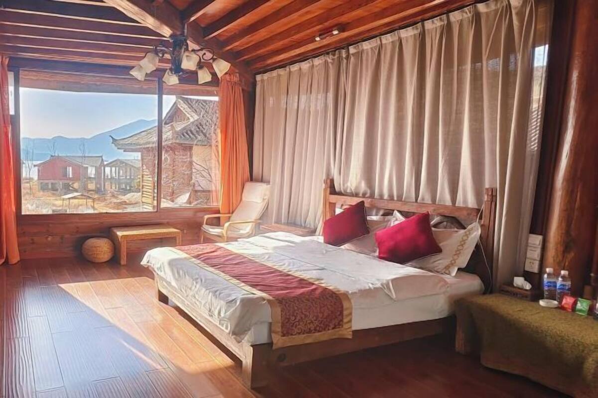 A wooden-framed bedroom with a bed, a chair, and a view of traditional buildings through a large window.