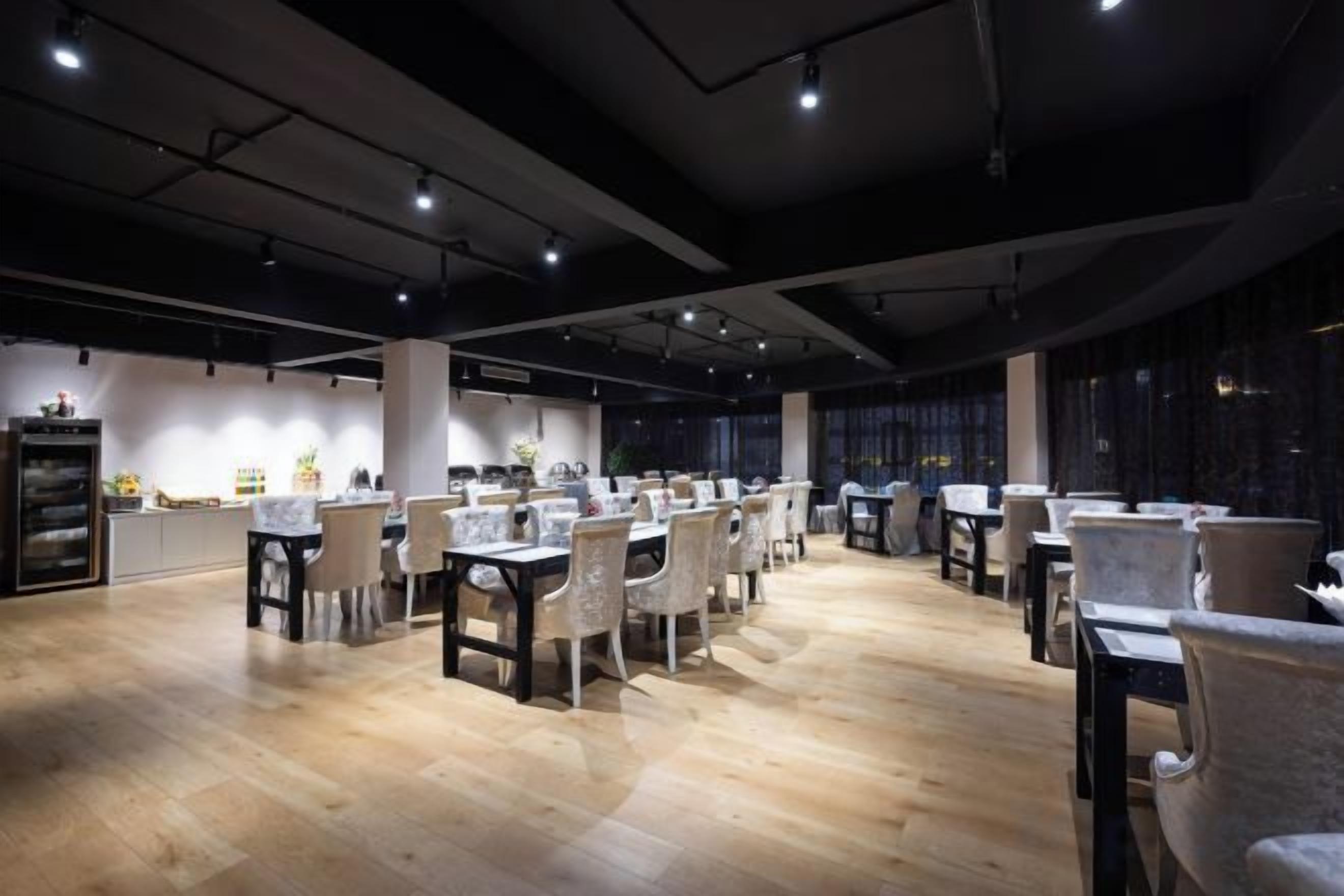 Uma ampla sala de jantar com várias mesas e cadeiras, uma cozinha ao fundo e um teto alto com vigas aparentes.