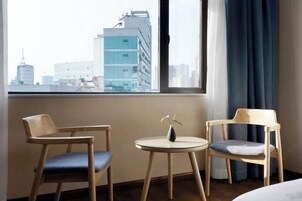 Quarto de hotel com uma janela grande, duas cadeiras de madeira, uma mesa redonda pequena e um vaso com flores.
