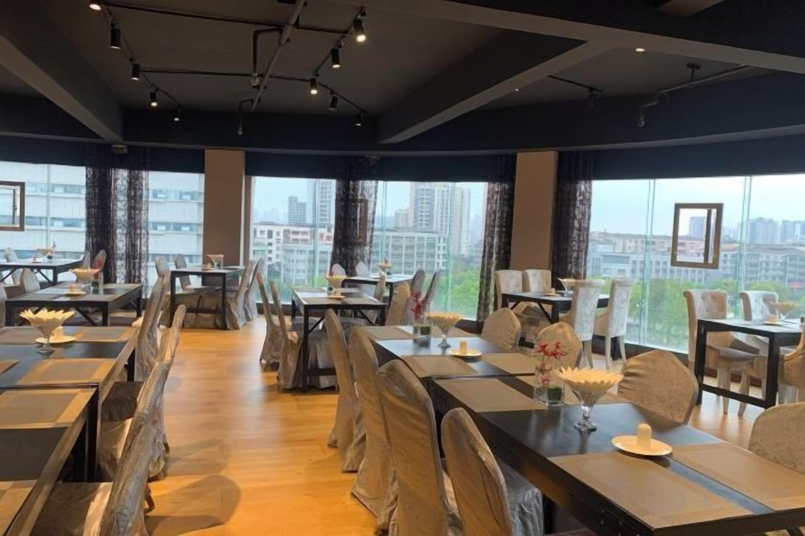 A modern dining area with multiple tables and chairs, large windows with a city view, and a high ceiling with exposed beams.
