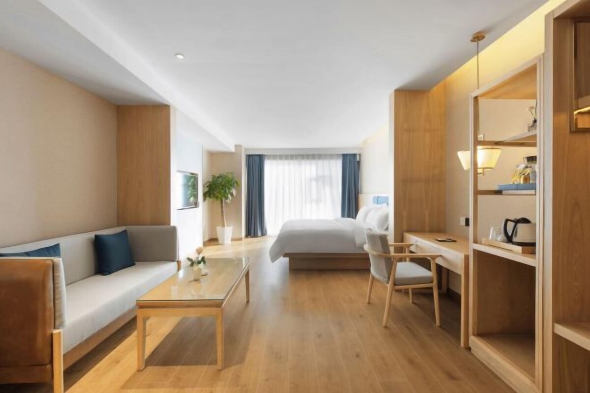 A modern hotel room with a wooden floor, a sofa, a bed, a desk, and a bookshelf.