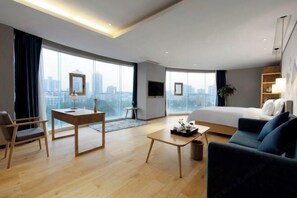 A modern hotel room with a large bed, a sofa, a desk, and a chair. There is a large window with a city view.