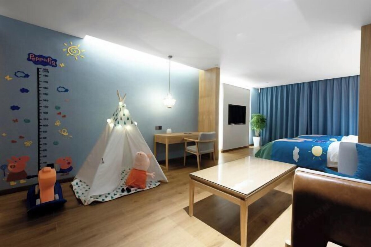 A child-friendly hotel room with a play tent, a desk, and a bed with a blue sky-themed bedspread.