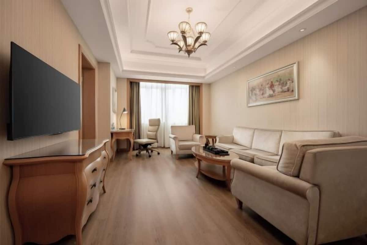 A hotel room with a flat-screen TV, a sofa, a coffee table, a desk, and a chandelier.