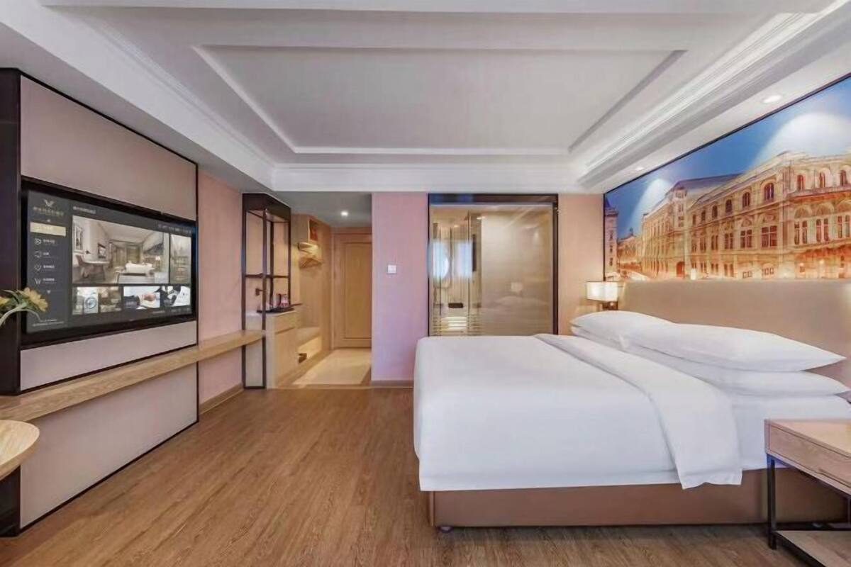 A hotel room with a large bed, a flat-screen TV mounted on the wall, and a mural of a building on the wall.