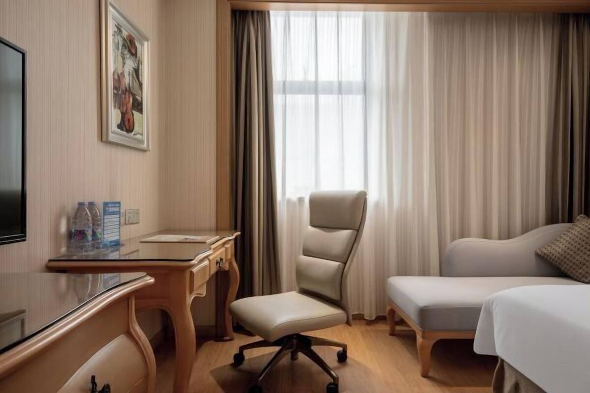 A hotel room with a desk, chair, bed, and a large window with curtains.