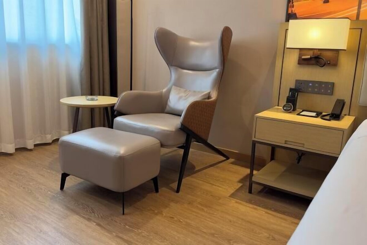 Una habitación de hotel moderna con un sillón de cuero, una mesita redonda pequeña, una mesilla de noche con una lámpara y una cama parcialmente visible.