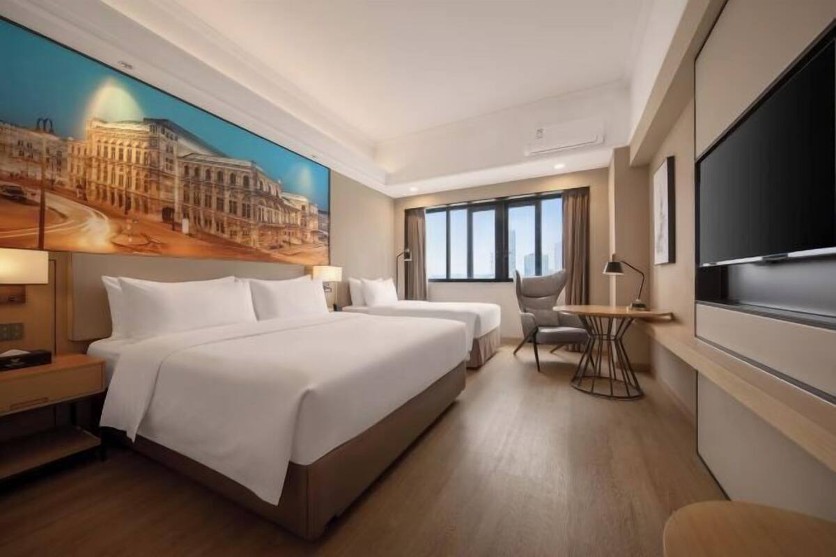 A hotel room with two beds, a desk, a chair, a TV, and a large window.