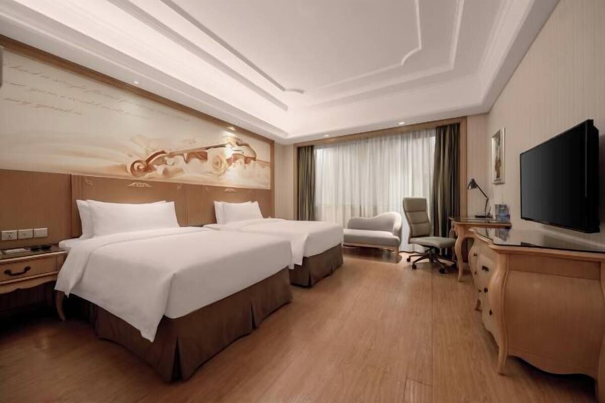 A hotel room with two beds, a desk, a TV, and a large window with curtains.