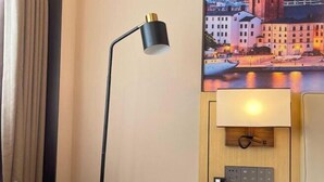 A hotel room with a bed, a desk with a lamp, a wall-mounted phone, and a painting of a cityscape. - Vienna International Hotel Foshan Qiandenghu Guicheng Subway Station Branch (Foshan)