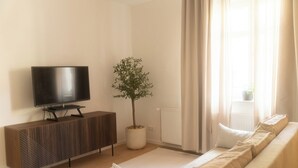 Una sala moderna con una televisión de pantalla plana sobre una consola de madera, una planta en maceta, un radiador y un sofá con cojines.