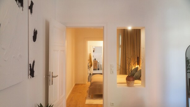 Un pasillo que conduce a un dormitorio con una cama, una mesita de noche y una planta en maceta.
