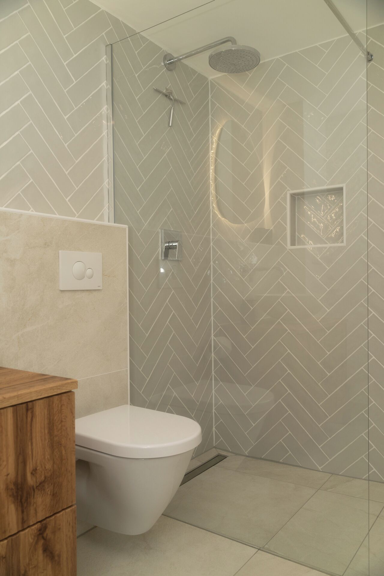 A modern bathroom with a herringbone tile pattern, a glass shower enclosure, a toilet, and a wooden vanity.