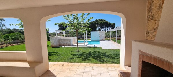 Une vue à travers une porte en arc donnant sur une cour arrière avec une piscine, une construction en pierre et un arbre.