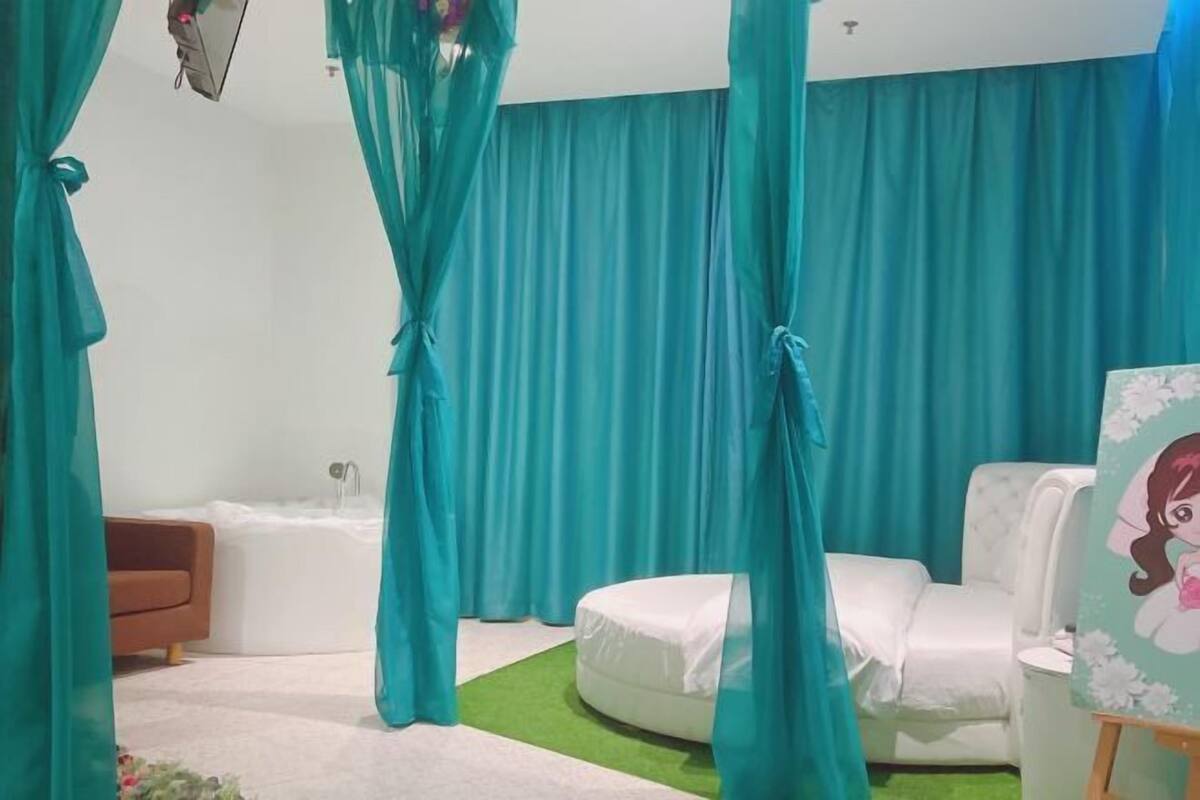 A room with teal curtains, a white bed, a white chair, and a small table with a painting.
