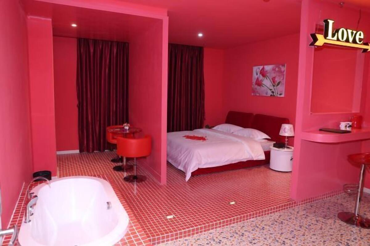 A pink-themed hotel room with a bed, a red chair, a white bathtub, and a vanity with a mirror.