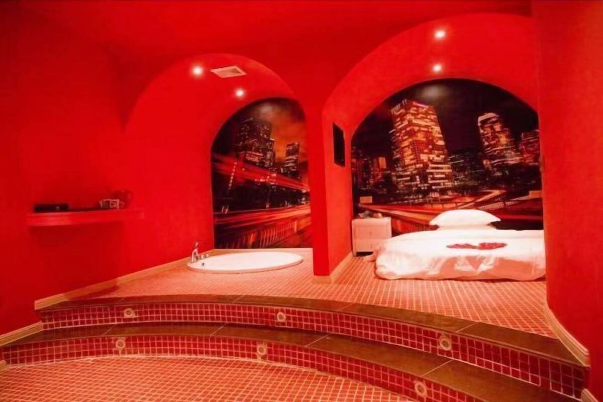 A red-themed room with a bathtub, a bed, and a cityscape mural.