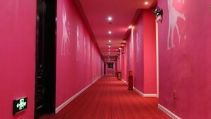 Un largo pasillo con alfombra roja y paredes rosadas, con una puerta a la izquierda que tiene un letrero verde.