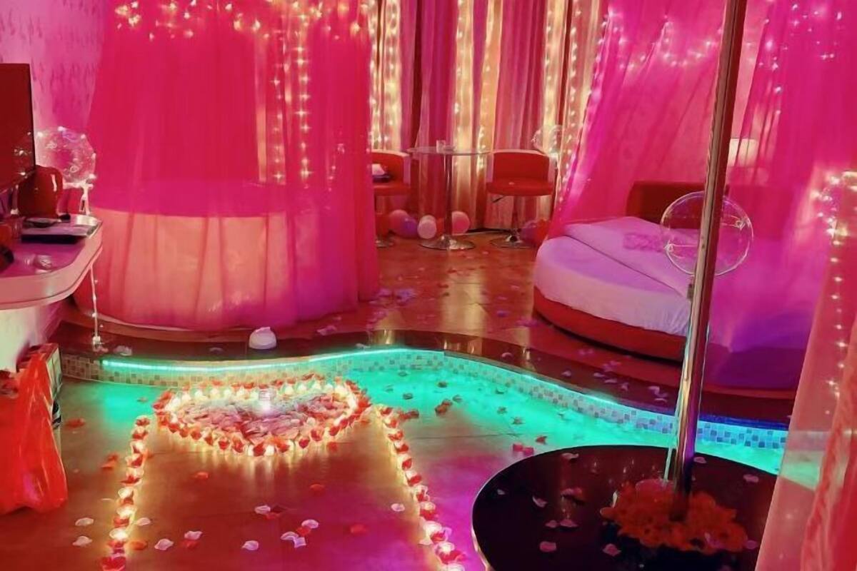 A room with a heart-shaped arrangement of rose petals, a bed, a chair, and a table with a glass.
