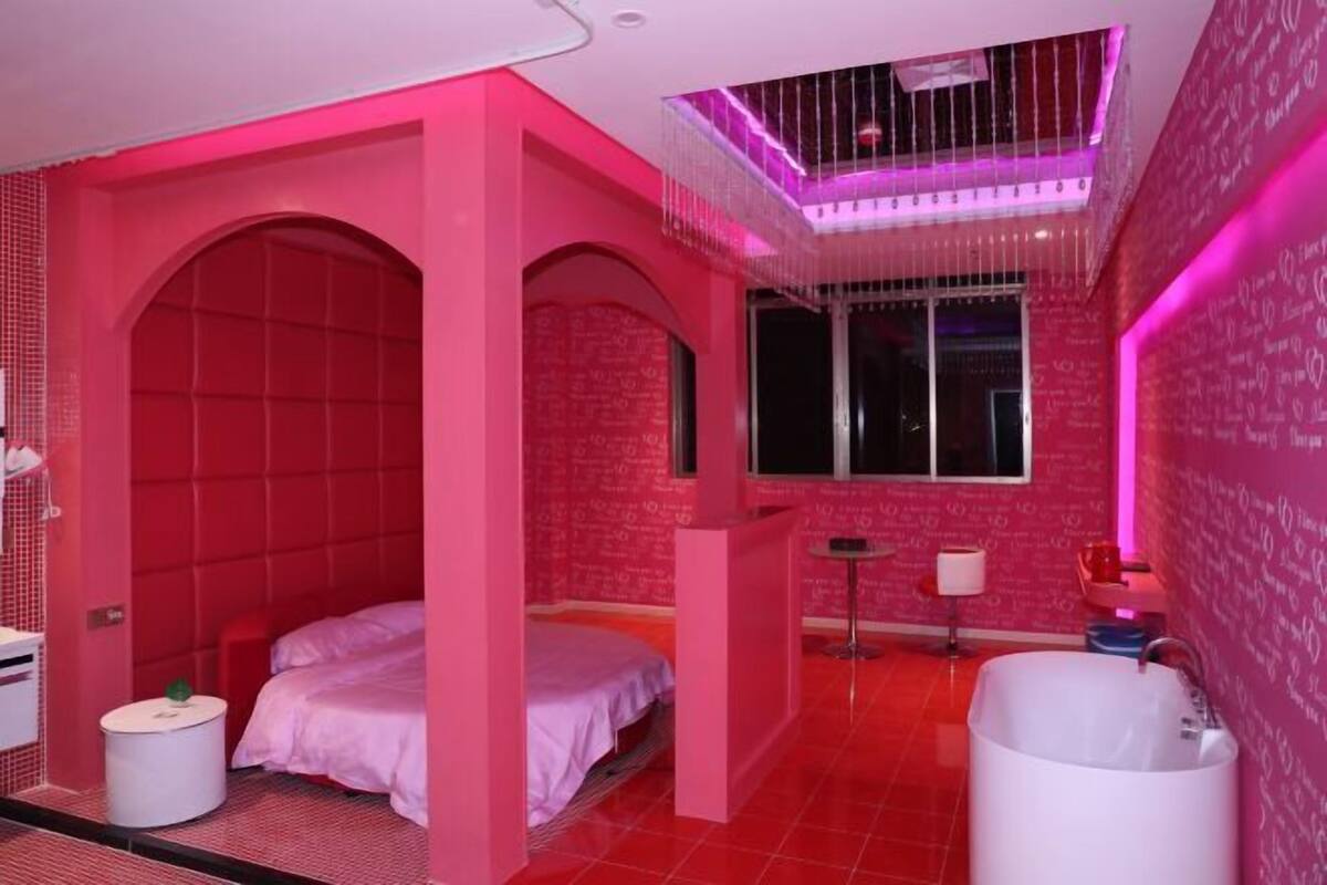 A pink-themed hotel room with a bed, a small round table, a white bathtub, and a window.