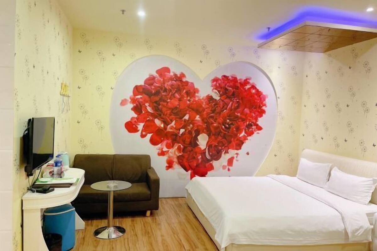 A hotel room with a bed, a sofa, a small table, a TV, and a wall with a heart-shaped floral design.