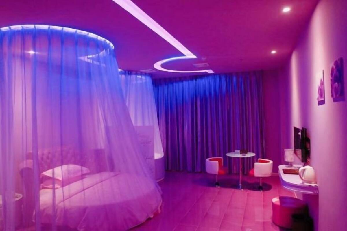 A room with a bed under a sheer canopy, a small round table, a chair, and a wall-mounted TV.