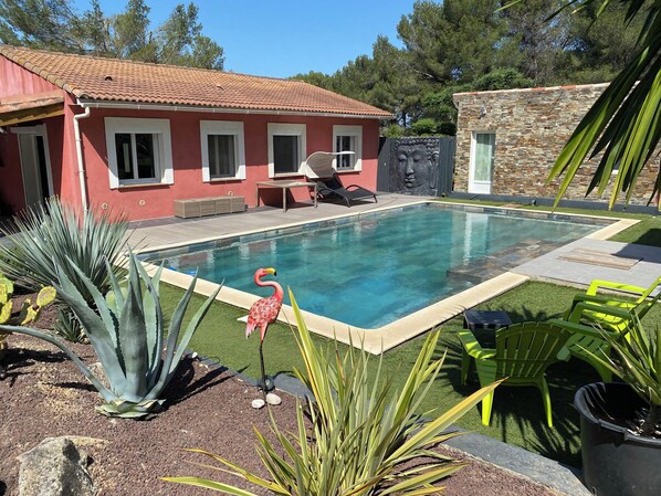 A swimming pool with a red house in the background, surrounded by plants and outdoor furniture.