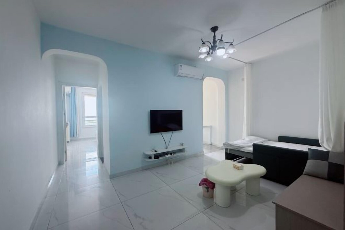 A modern living room with a flat-screen TV, a seating area with a bench and ottoman, and a dining table.