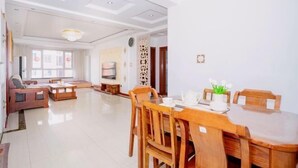 A spacious living room with a wooden dining table, chairs, and a sofa. There is a television and a decorative wall panel. - Qingdao Yijian Qingxin Whole Vision Apartment (Qingdao)