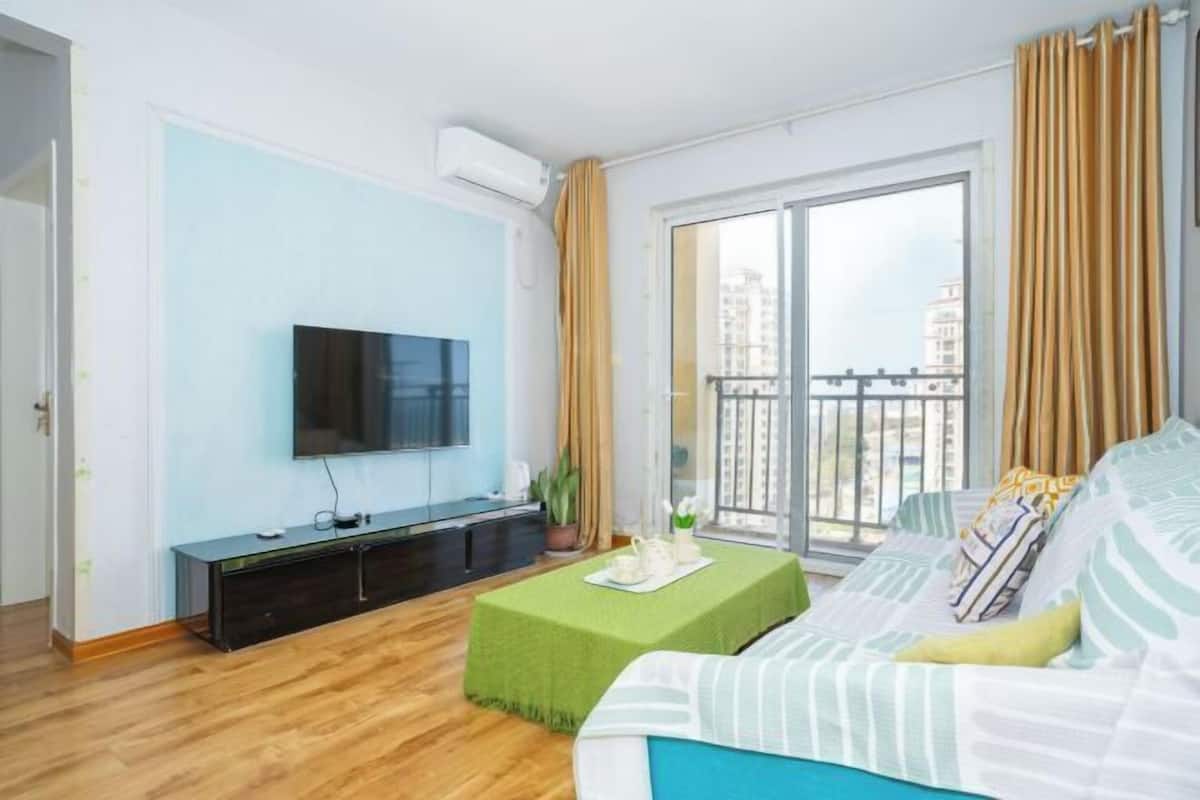 A modern living room with a flat-screen TV, a sofa, a coffee table, and a balcony with curtains.