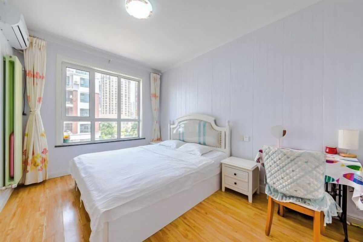 A bedroom with a bed, a bedside table, a chair, a window, and a desk with a lamp.