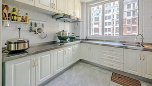 A kitchen with white cabinets, a window with a view of buildings, and a countertop with a rice cooker and pots. - Qingdao Yijian Qingxin Whole Vision Apartment (Qingdao)