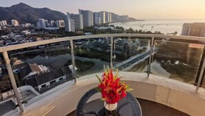 Un balcon doté d’une balustrade en verre, un vase rempli de fleurs rouges et une vue sur un paysage urbain avec des bâtiments et une étendue d’eau.