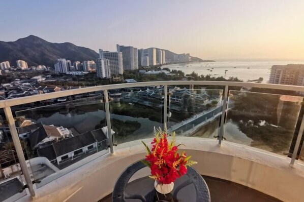 A balcony with a glass railing, a vase with red flowers, and a view of a cityscape with buildings and a body of water.