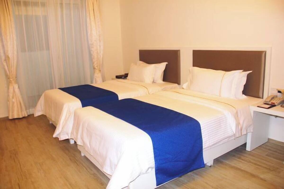 Ein Hotelzimmer mit zwei Betten, einem Nachttisch mit Telefon und einem Fenster mit leichtem Vorhang.