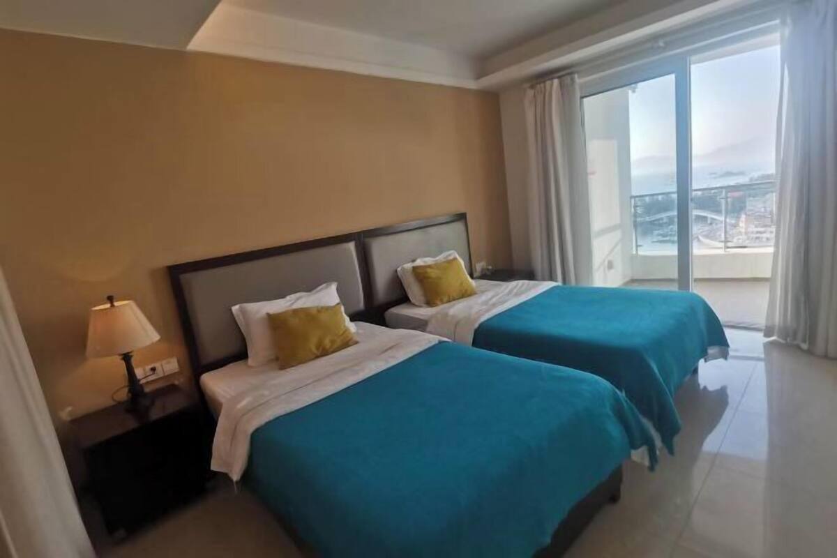 Ein Hotelzimmer mit zwei Betten, einem Nachttisch mit Lampe und Blick auf eine Stadtlandschaft durch ein großes Fenster.