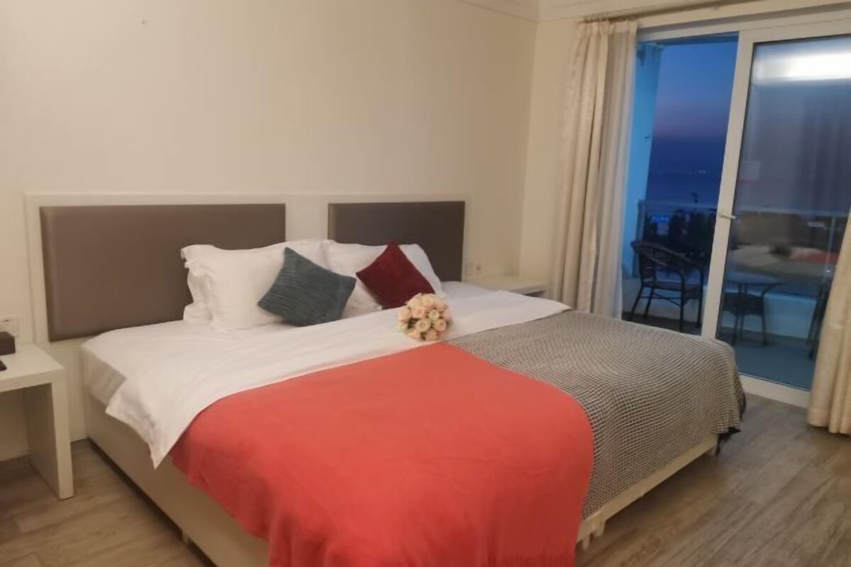 A neatly made bed with a red blanket and a bouquet of flowers, a white nightstand, and a view of a balcony with a chair.