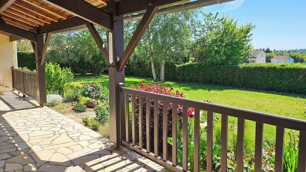 A covered outdoor area with a stone floor and wooden railing, overlooking a well-maintained garden and a distant building. - Villa de Charme Avec Piscine - Idéale Pour des Vacances en Famille ou Entre Amis (Julienne)