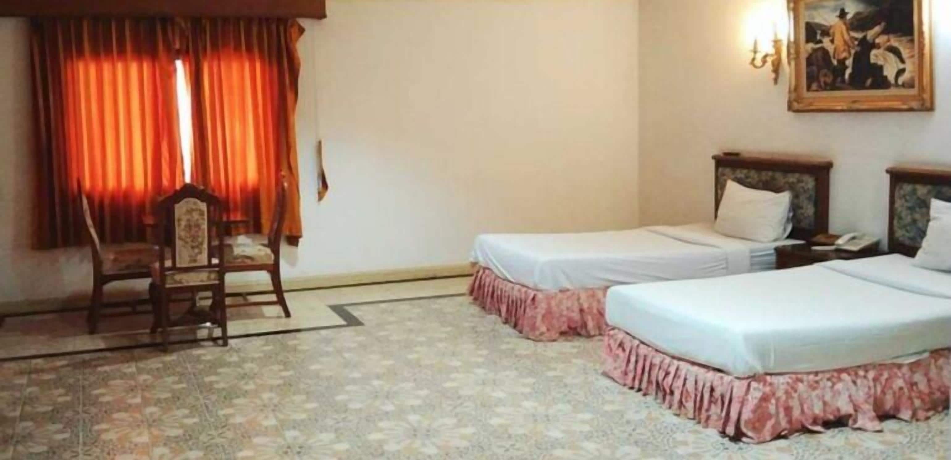 A hotel room with two beds, a chair, a painting on the wall, and a window with curtains.