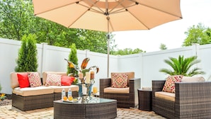 An outdoor patio area with wicker furniture, a glass coffee table, and a large umbrella.
