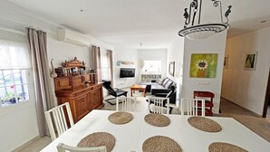 A modern living room with a dining area, featuring a white table with woven placemats, a sofa, a TV, and a chandelier.