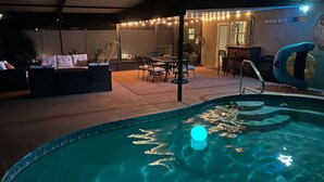 Une piscine avec une lumière bleue flottant à la surface, entourée de sièges d’extérieur et illuminée de guirlandes lumineuses.
