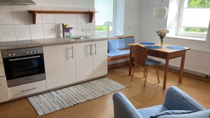 Moderni keittiö, jossa on valkoiset kaapit, Bosch-uuni, puinen pöytä sinisillä tyynyillä ja ikkuna, josta avautuu näkymä vehreään maisemaan.