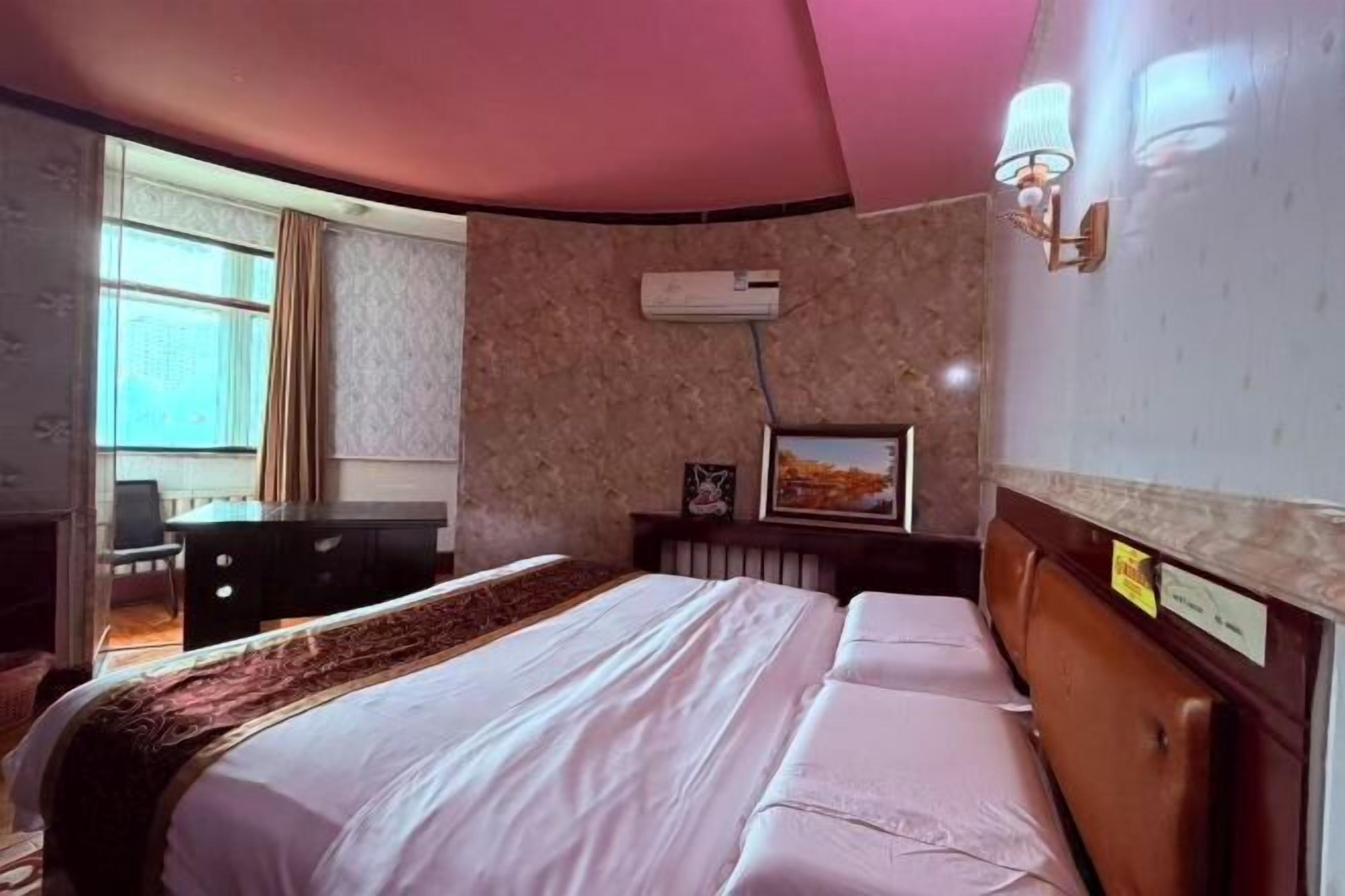 A hotel room with a bed, a desk, an air conditioner, a window, and a wall-mounted lamp.