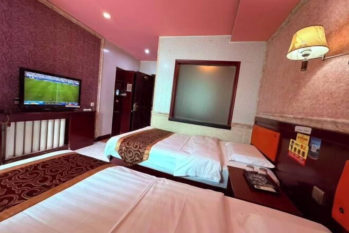 Quarto de hotel com duas camas, televisão, uma janela grande e uma luminária fixada na parede.