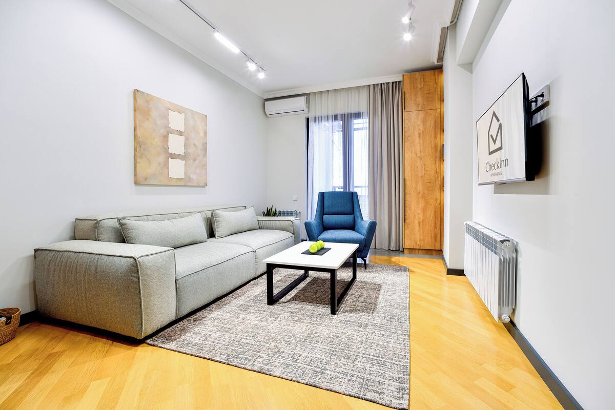 Un salon moderne avec un canapé gris, un fauteuil bleu, une table basse sur laquelle se trouvent des pommes, un tableau accroché au mur et une enseigne « Checkinn ».