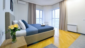 Ein modernes Hotelzimmer mit einem großen Bett, einem Nachttisch mit Blumenvase und Blick auf die Stadt durch große Fenster.